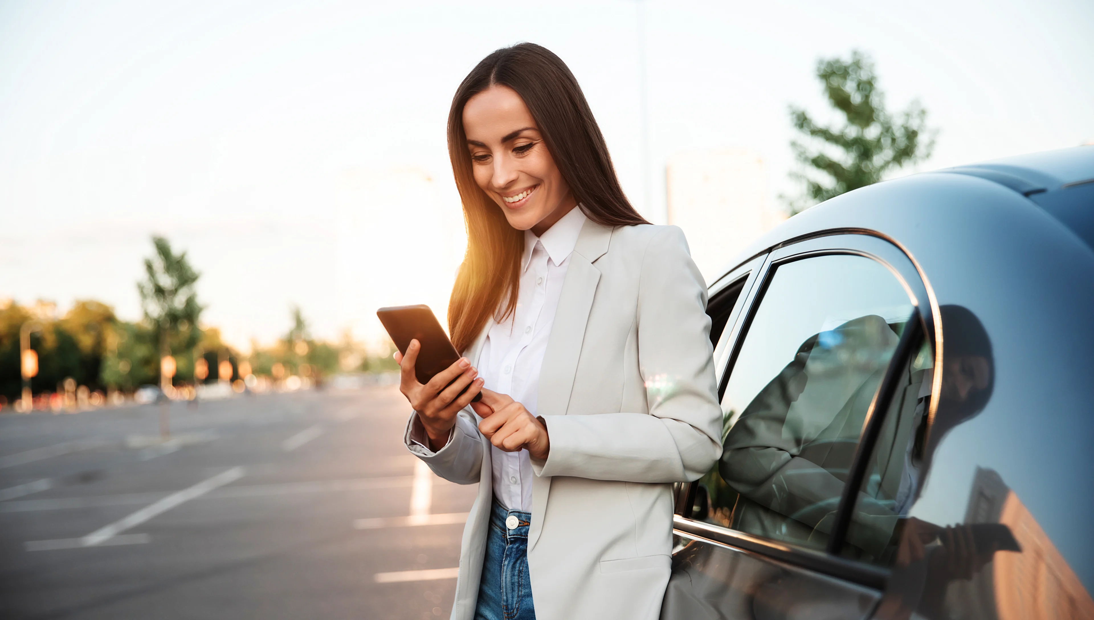 A woman in a light blazer stands by a parked car, smiling while looking at her smartphone in a parking lot during daylight. Trees and buildings are visible in the blurred background.