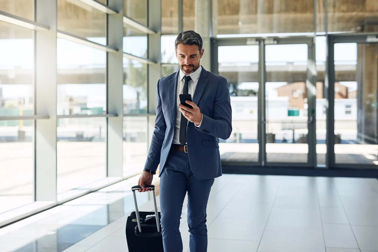 Business traveller walking through an airport terminal while looking at his phone and pulling a suitcase.